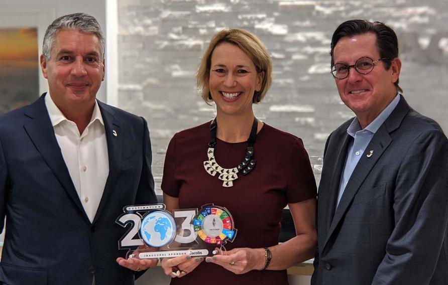 Two white men flanking a blond woman in a maroon dress and statement necklace holding a 2030 trophy